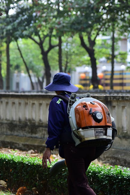 A person wearing a dark long-sleeved shirt and a grey cap is trimming a neatly maintained hedge with hedge shears. The hedge has dense, green foliage with some leaves showing a hint of new growth. Beh